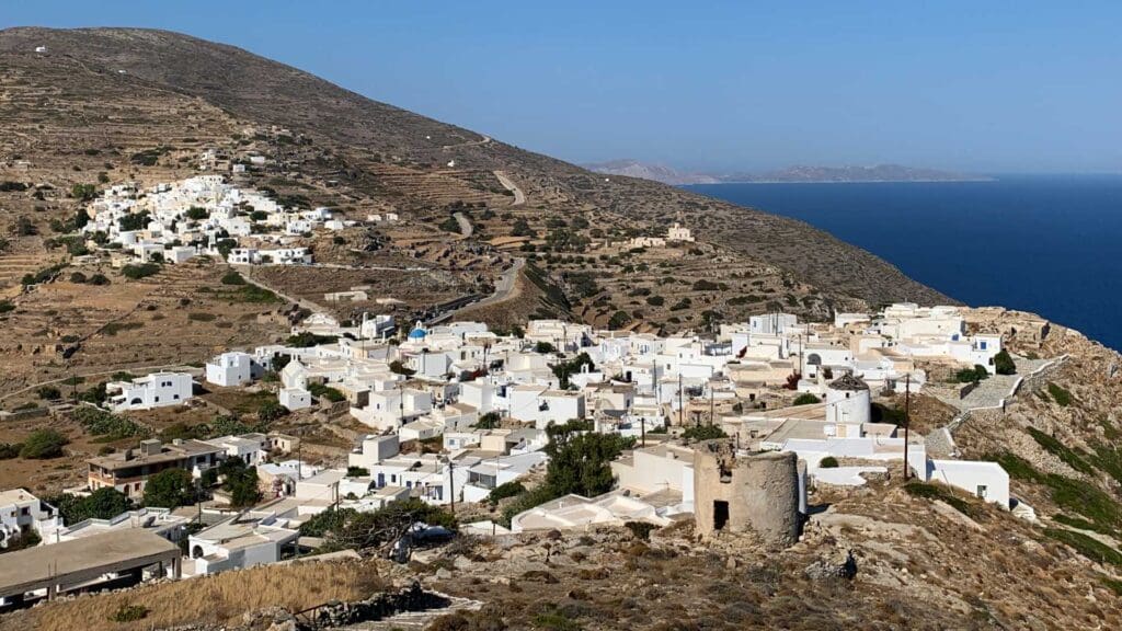 Aerial view of Sikinos Island's coastal village, with white buildings perched on a hillside, winding roads meandering through, and the shimmering sea in the background.