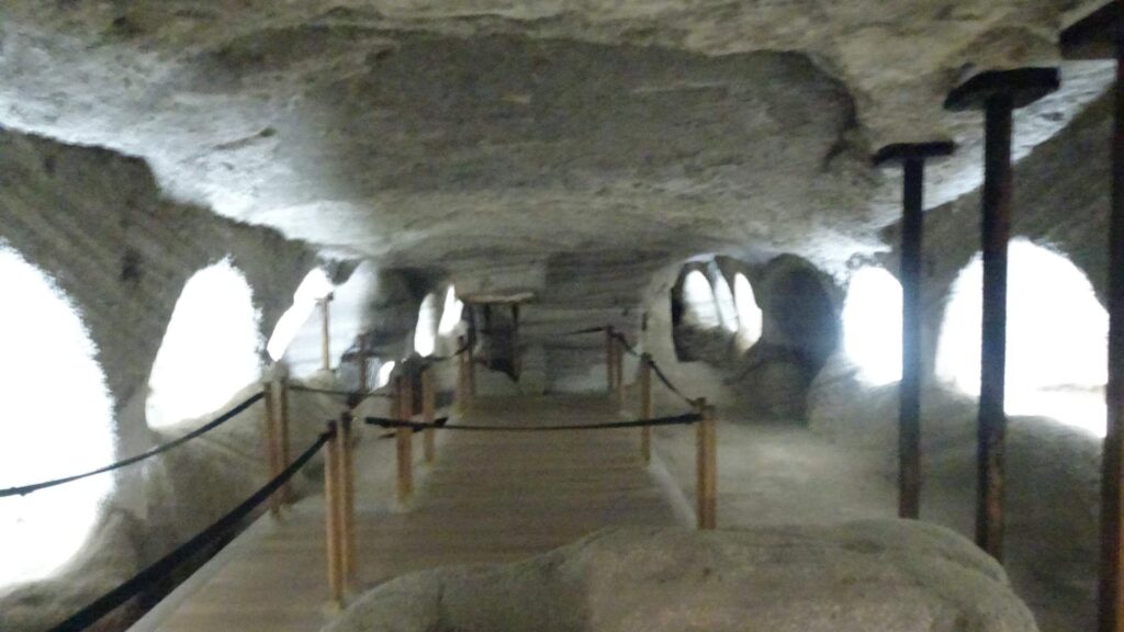 A dimly lit underground cave corridor on Milos features stone walls, arched openings, and wooden railings along a narrow walkway.