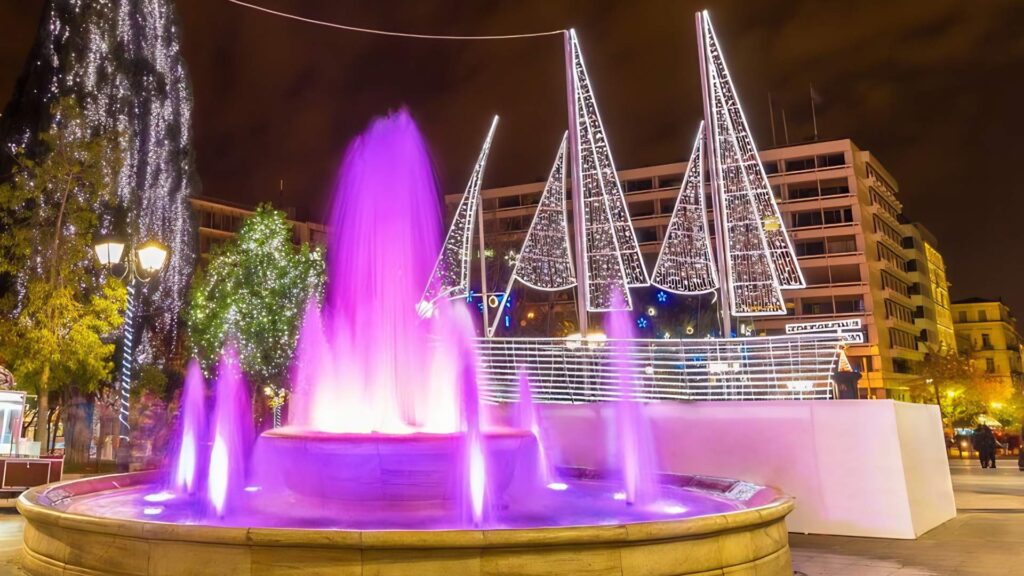 A vibrant purple fountain is illuminated at night, surrounded by buildings and trees. In the background, decorative sail-shaped lights are displayed, creating a magical atmosphere perfect for enjoying Christmas in Athens 2025.