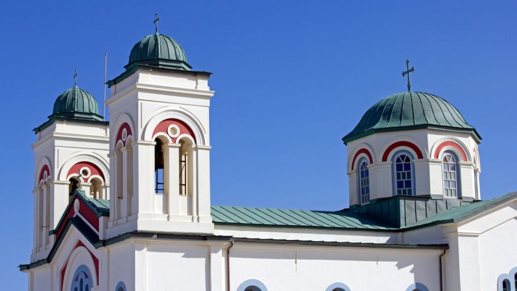 The white church in Naoussa, Paros, stands gracefully with two tall bell towers and a dome, sporting green roofs and red arch decorations, all set against a clear blue sky.