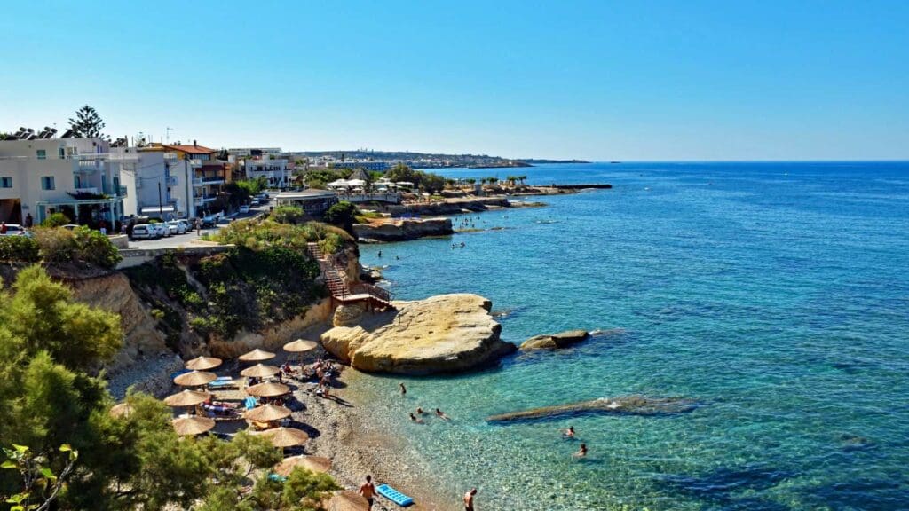 Coastal scene with clear blue water, rocky shores, and beachgoers swimming at Heraklion beaches; sunbathers relax under umbrellas as buildings and greenery line the shoreline under a clear sky.
