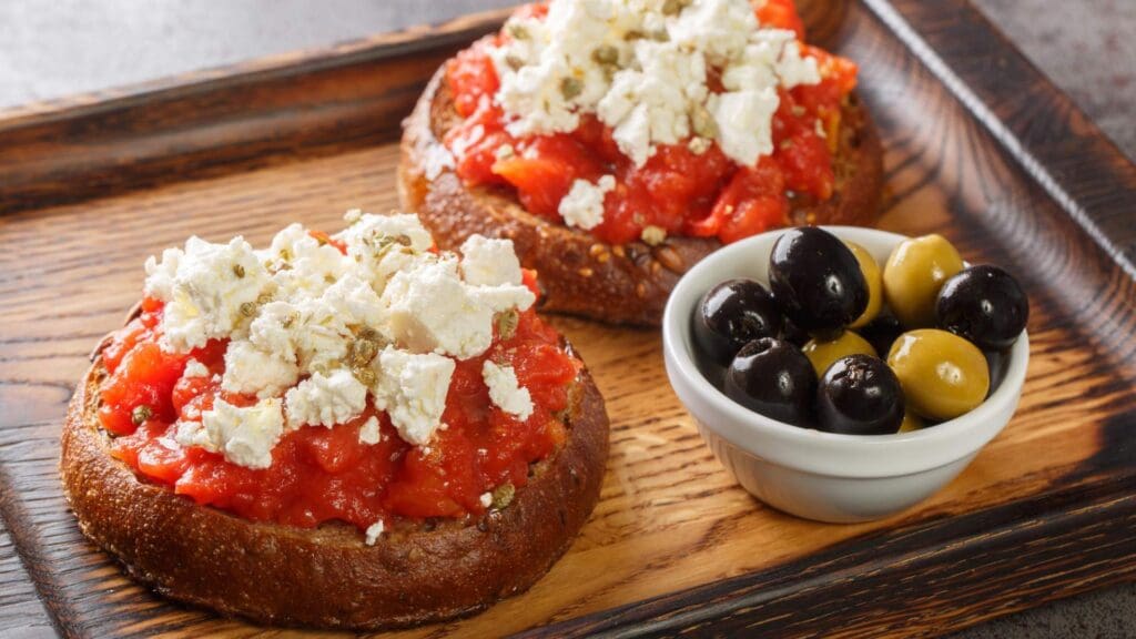 Two slices of bread topped with tomatoes and cheese rest on a wooden tray, reminiscent of Greek street food, accompanied by a small bowl of black and green olives.