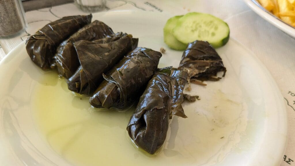 A plate featuring six grape leaf rolls, partially filled with rice, and two cucumber slices, drizzled with olive oil—capturing the essence of Greek street food.
