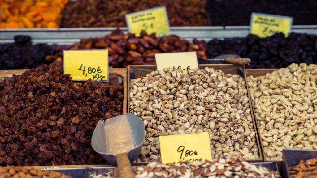 A market stall with assorted nuts and dried fruits reminiscent of Greek street food. Pistachios and cashews are displayed with price tags in euros, while a metal scoop rests among raisins.