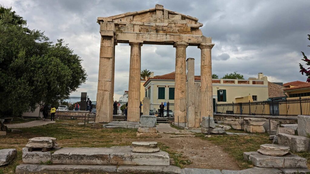 Explore the ruins of a stone gateway with columns at an archaeological site, reminiscent of the magnificent Acropolis, surrounded by trees and buildings under a cloudy sky.