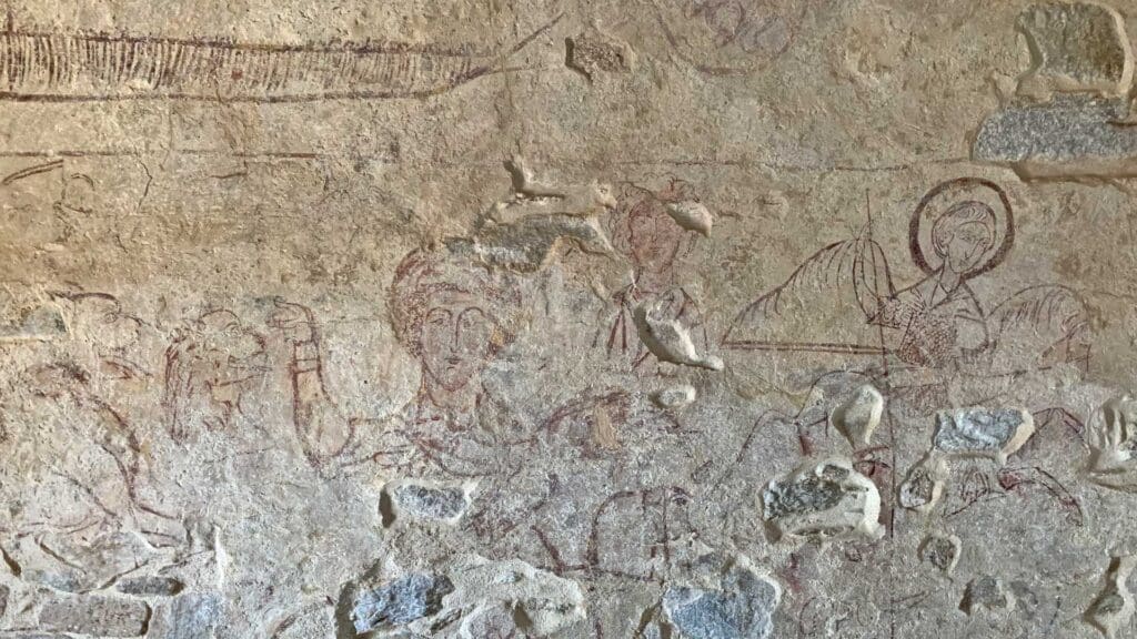 A faded ancient wall painting on Sikinos Island captures human figures and elements, some adorned with halos, set against a backdrop of deteriorated stone.