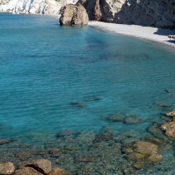 A rocky beach on the picturesque Milos Beaches with clear blue water, lined with lounge chairs and umbrellas on the right. Cliffs rise majestically in the background under a serene blue sky—a must-see spot for any traveler.