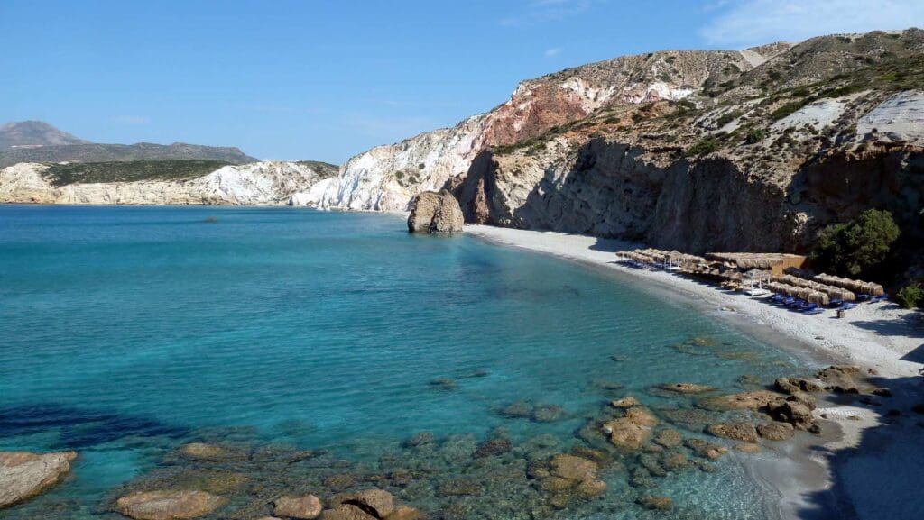 Discover Milos, where a clear blue sea meets rocky cliffs and a quaint beach lined with umbrellas and sun loungers. Sparse greenery dots the coast under a clear sky, making it one of the must-see spots among Milos beaches.