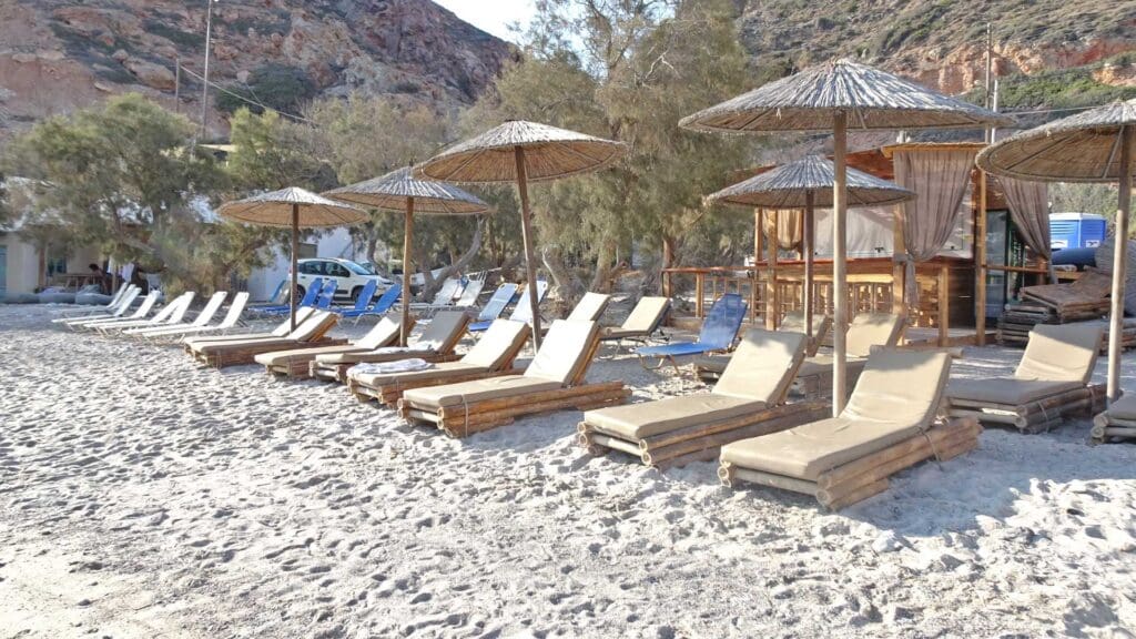 A row of sun loungers with straw umbrellas adorns the sandy shores of one of Milos Beaches, framed by trees and rocky hills in the background—a must-see spot for those looking to discover Milos.