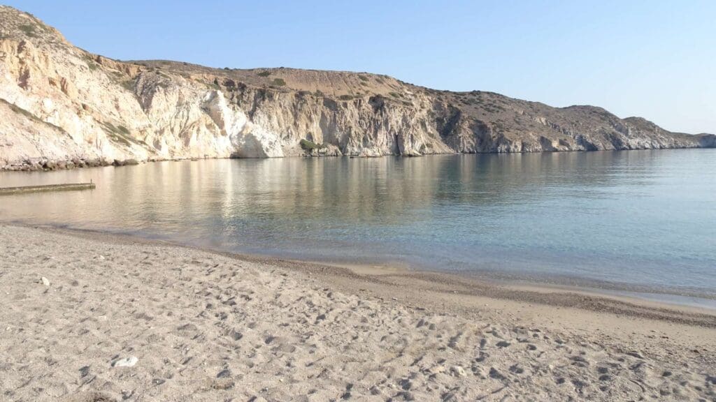 Discover Milos as a calm, clear sea laps against a sandy beach, with rugged cliffs in the background under a clear blue sky. This is one of the must-see spots among the beautiful Milos beaches.