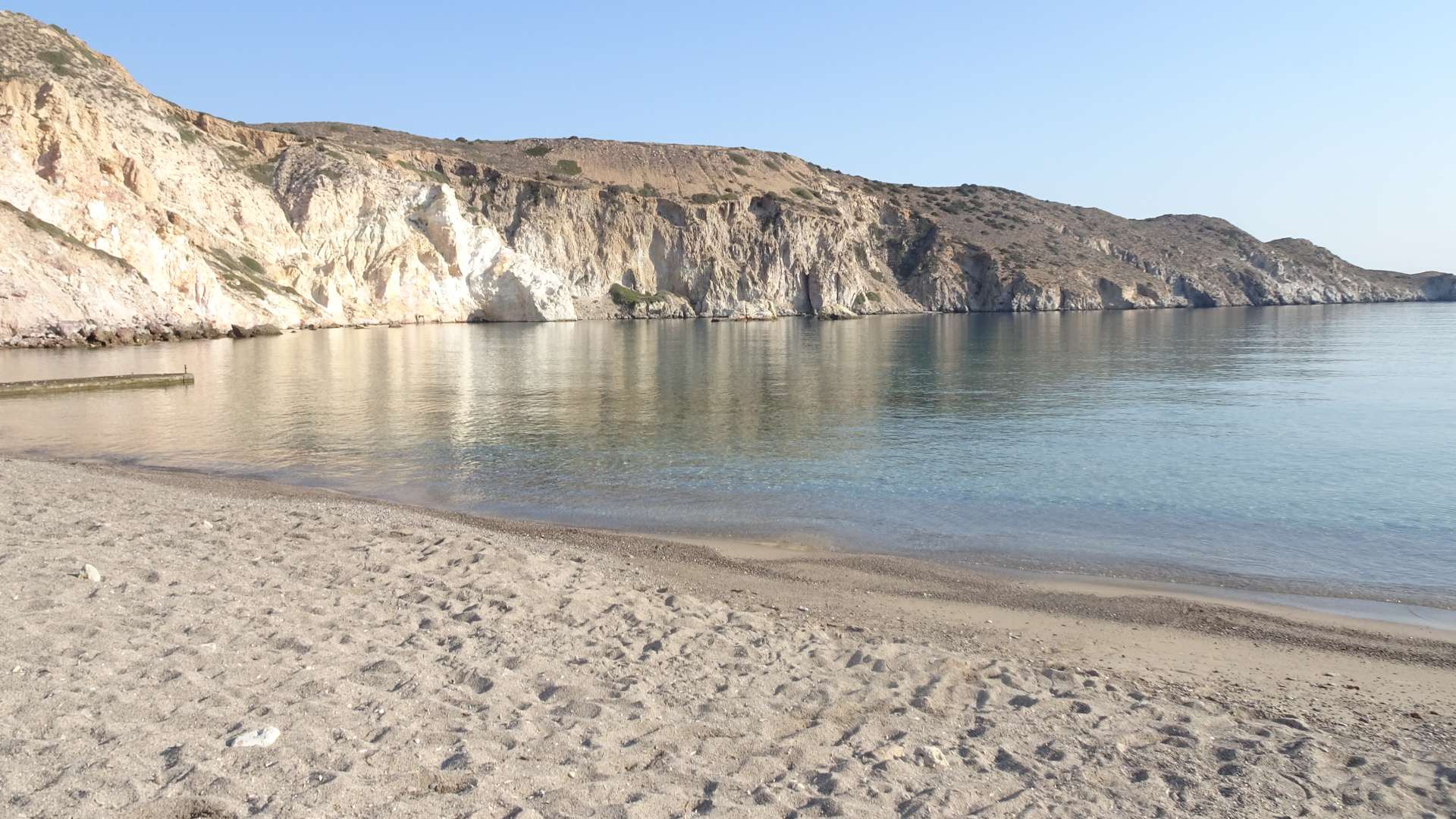 Discover Milos as a calm, clear sea laps against a sandy beach, with rugged cliffs in the background under a clear blue sky. This is one of the must-see spots among the beautiful Milos beaches.