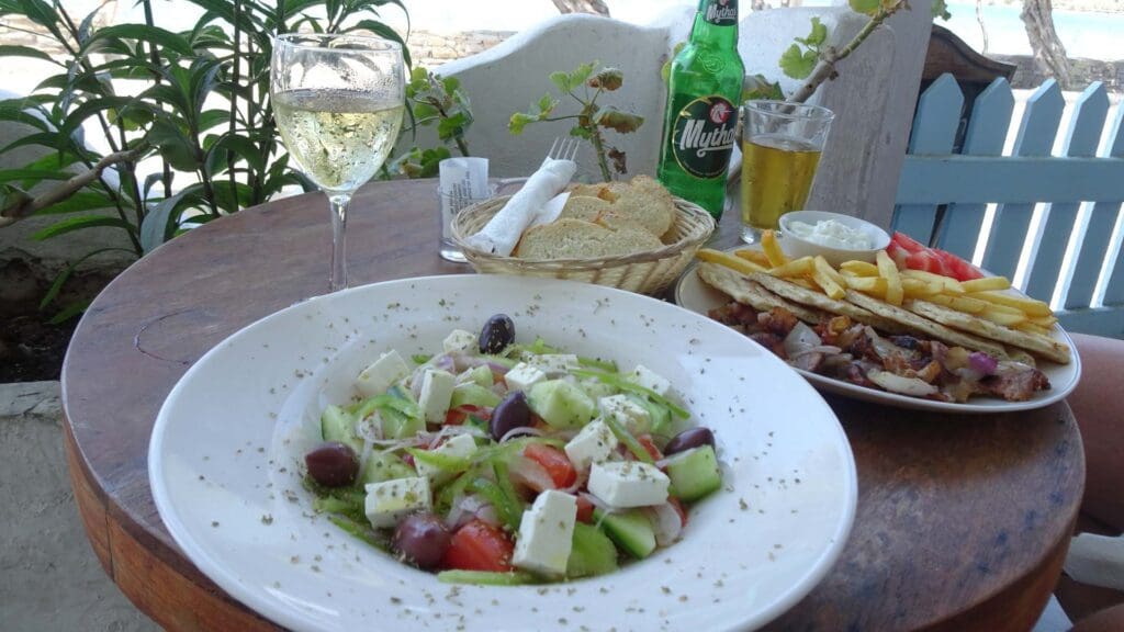 A table with a Greek salad, grilled meat platter reminiscent of Greek street food, a bread basket, a glass of white wine, and a bottle of Mythos beer. There's a seaside view in the background.