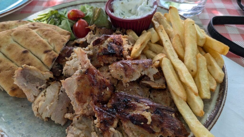 A plate of grilled meat with fries, pita bread, and salad, served with a small dish of sauce on a checkered tablecloth captures the essence of Greek street food.