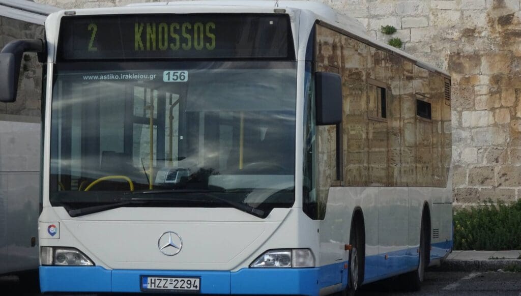 A white and blue bus labeled "2 Knossos" is parked in front of a stone wall, ready to whisk travelers away to the nearby Heraklion beaches.