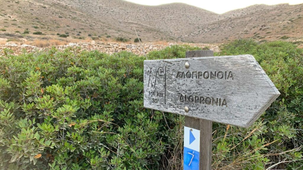 A rustic wooden trail sign surrounded by green bushes points towards a rocky mountain path, evoking the charm of Sikinos Island.