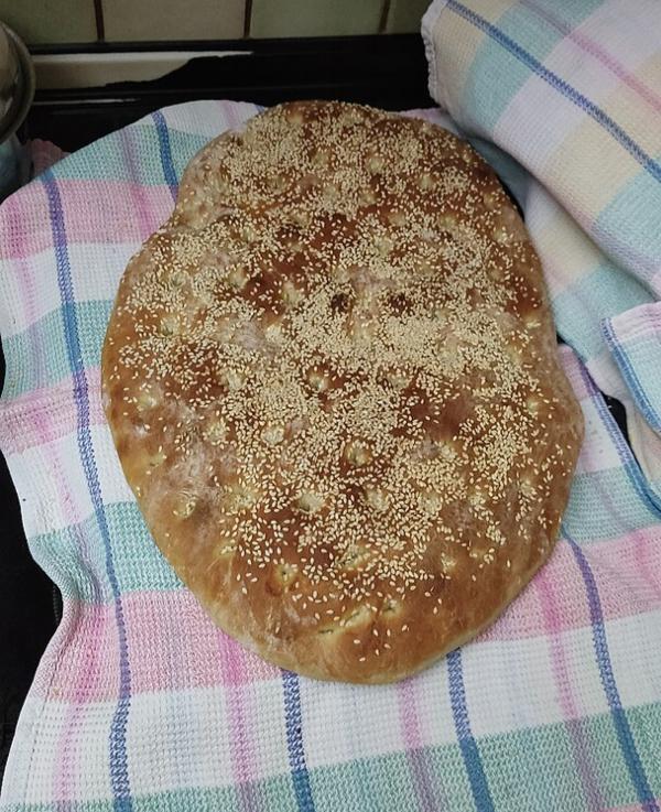 A large, round loaf of bread topped with sesame seeds rests on a plaid cloth, reminiscent of the festive spirit during Apokries.