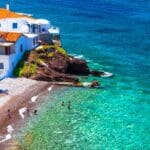 A coastal scene with white buildings and orange roofs overlooks Hydra beaches, where straw umbrellas line the shore and people swim in the clear turquoise water.