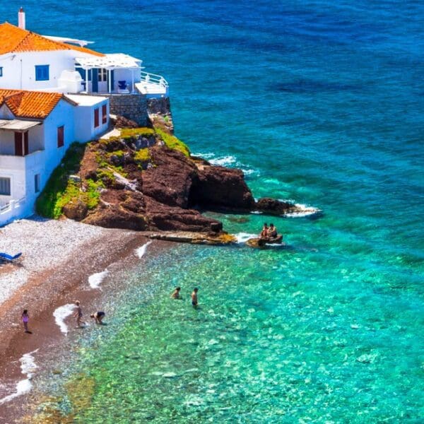 A coastal scene with white buildings and orange roofs overlooks Hydra beaches, where straw umbrellas line the shore and people swim in the clear turquoise water.