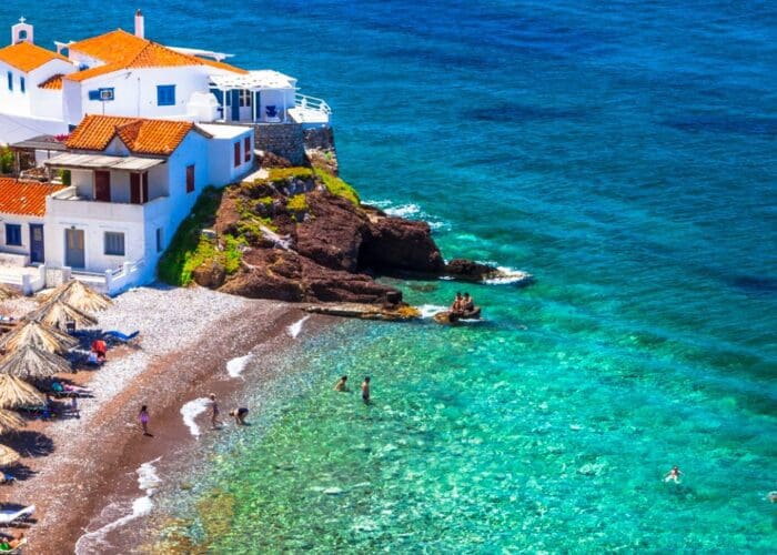 A coastal scene with white buildings and orange roofs overlooks Hydra beaches, where straw umbrellas line the shore and people swim in the clear turquoise water.