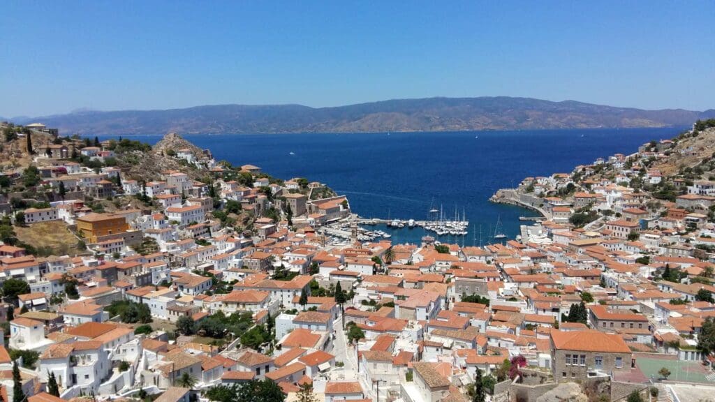 Aerial view of a coastal town with red-roofed buildings and a marina, surrounded by hills and the blue sea of Hydra beaches under a clear sky.