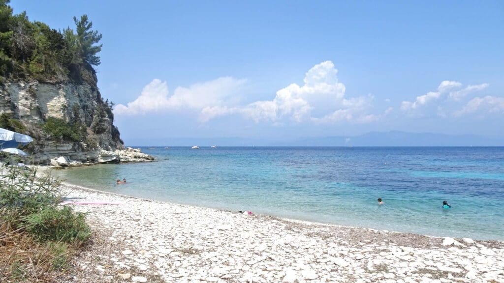 A Greek island-hopping traveler might find paradise at this rocky beach, where the clear blue water meets a towering cliff to the left. Under a sky adorned with scattered clouds, swimmers immerse themselves in the serene sea, crafting memories that any wanderer's itinerary would treasure.