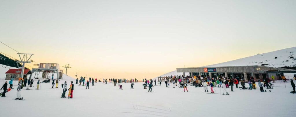 A busy ski resort with people in colorful ski gear gathered near ski lifts and a lodge at sunset, reminiscent of the vibrant scene at some of the best Greek ski resorts. Snow-covered ground and a clear sky enhance this splendid view.