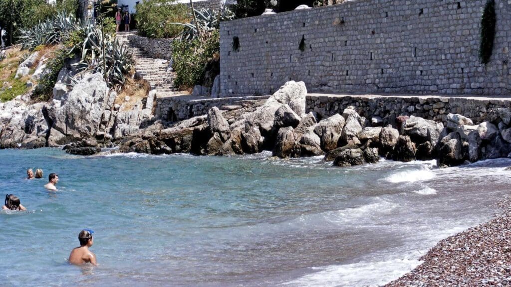 People swimming near a rocky beach with a stone wall and steps reminiscent of Hydra beaches, leading up to a lush garden area.