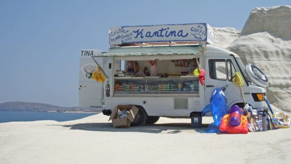 A mobile food truck at Sarakiniko Beach, Milos, offers refreshing cold drinks, iced coffee, and snacks to beachgoers. Vibrant inflatables add charm to the scene, creating a perfect stop in any visitor's guide.