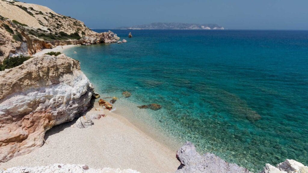 A rocky coastal beach with clear turquoise water and distant islands under a blue sky, Milos Beaches offer a stunning vista that's a must-see spot for any Milos travel itinerary.