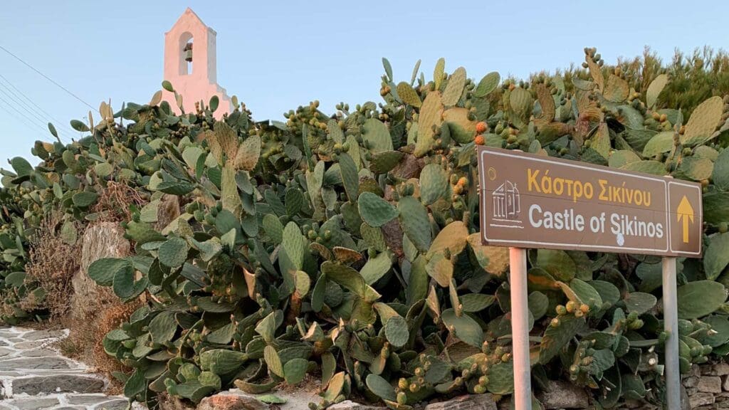 A sign reads "Castle of Sikinos" with an arrow, nestled among dense cactus plants on Sikinos Island. In the background, a small white church stands peacefully.