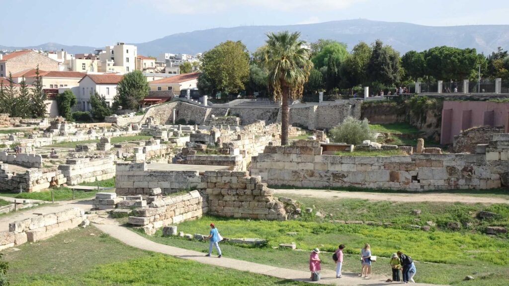 People explore the ancient ruins of the Acropolis, with distant mountains and modern buildings silhouetted against a clear sky. Expert tips guide their journey, offering historical insights that bring the past to life amid this timeless landscape.