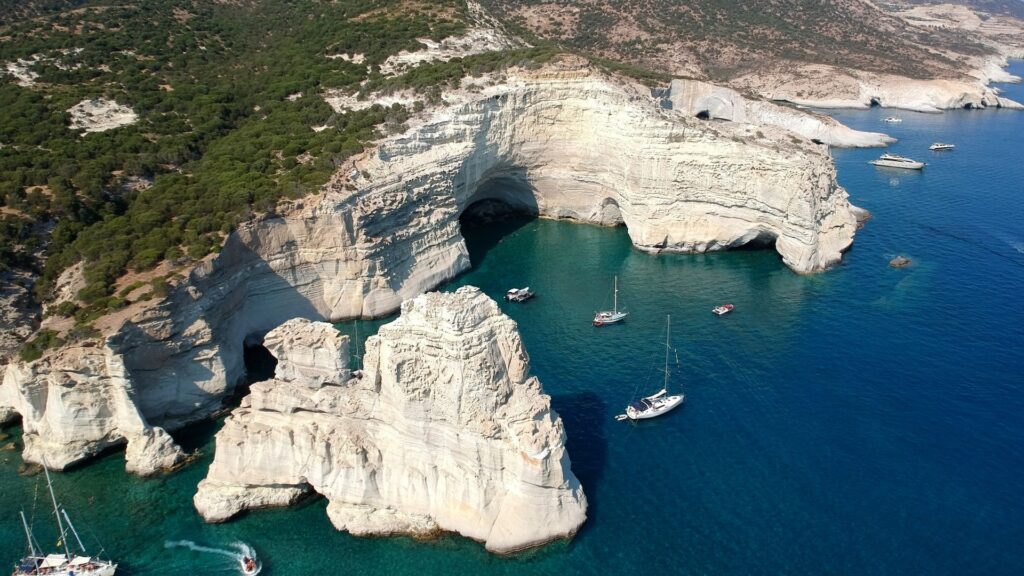 Aerial view of turquoise water and sailboats near the white rocky cliffs of Milos, adorned with lush greenery on top.