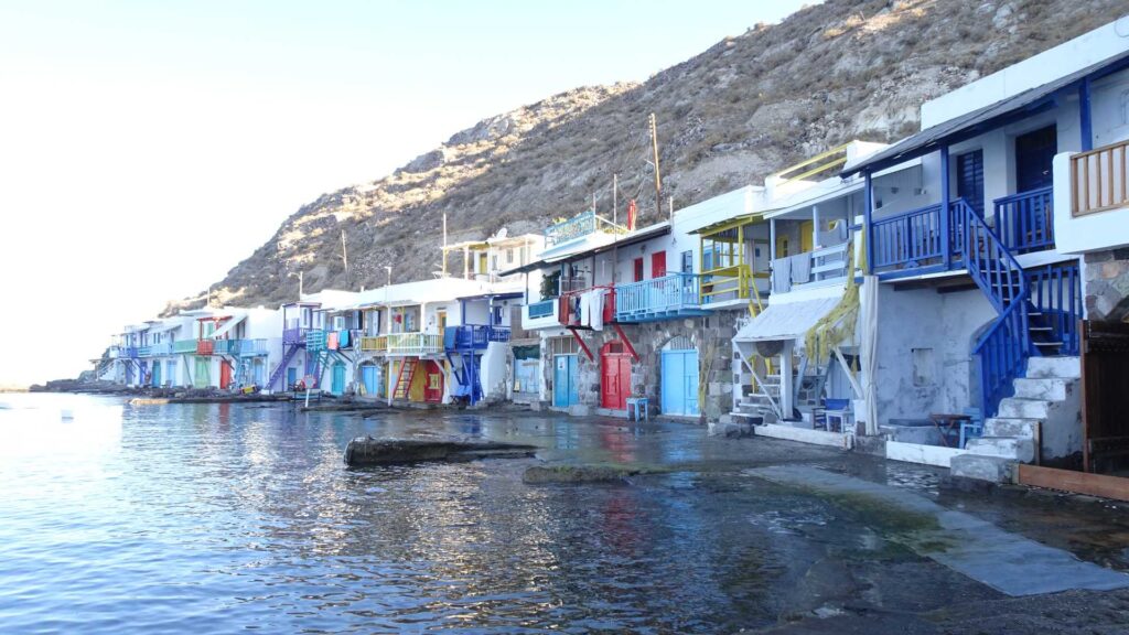 A row of colorful seaside houses with balconies overlooks the calm waters of Milos, set against a rocky hillside, offering a picture-perfect scene from any island guide.