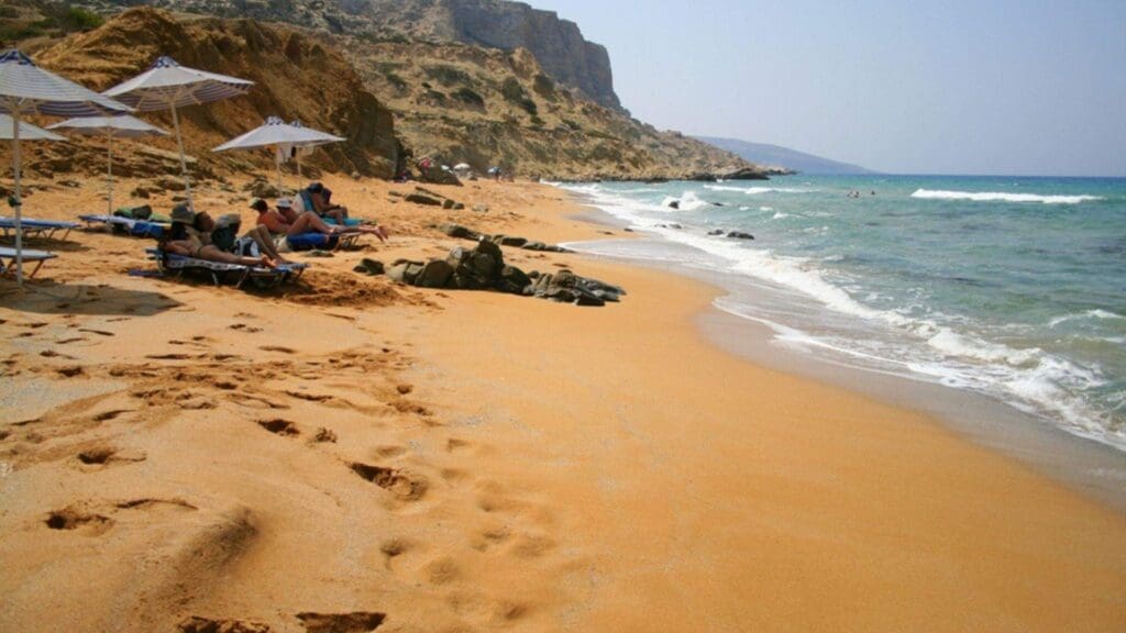 The sandy beach, a gem among Heraklion beaches, boasts soothing waves and majestic rocky cliffs. Visitors unwind on sun loungers under colorful umbrellas, capturing the essence of a perfect seaside escape.