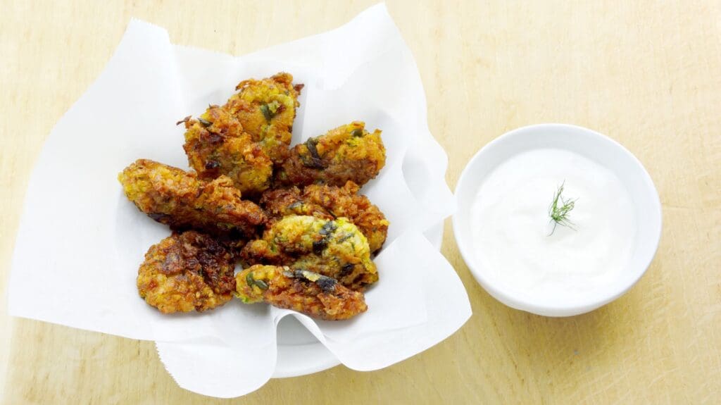 A plate of zucchini fritters, reminiscent of Greek street food, rests on a paper towel beside a small bowl of yogurt sauce garnished with a dill sprig.