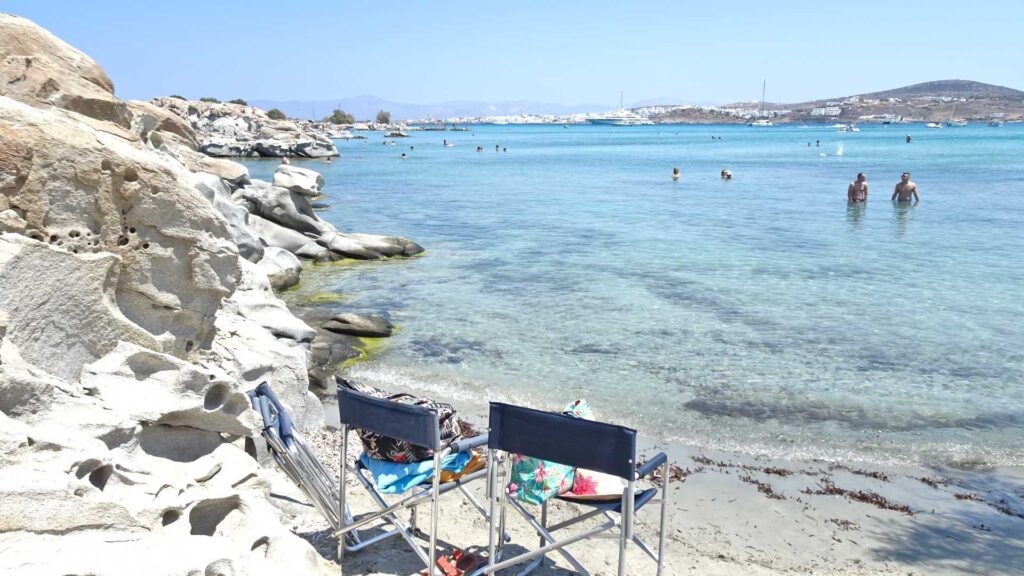 Two empty chairs on a sandy beach by clear blue water, reminiscent of the serene shores of Paros. People swim in the background near rocky outcrops under a clear sky.