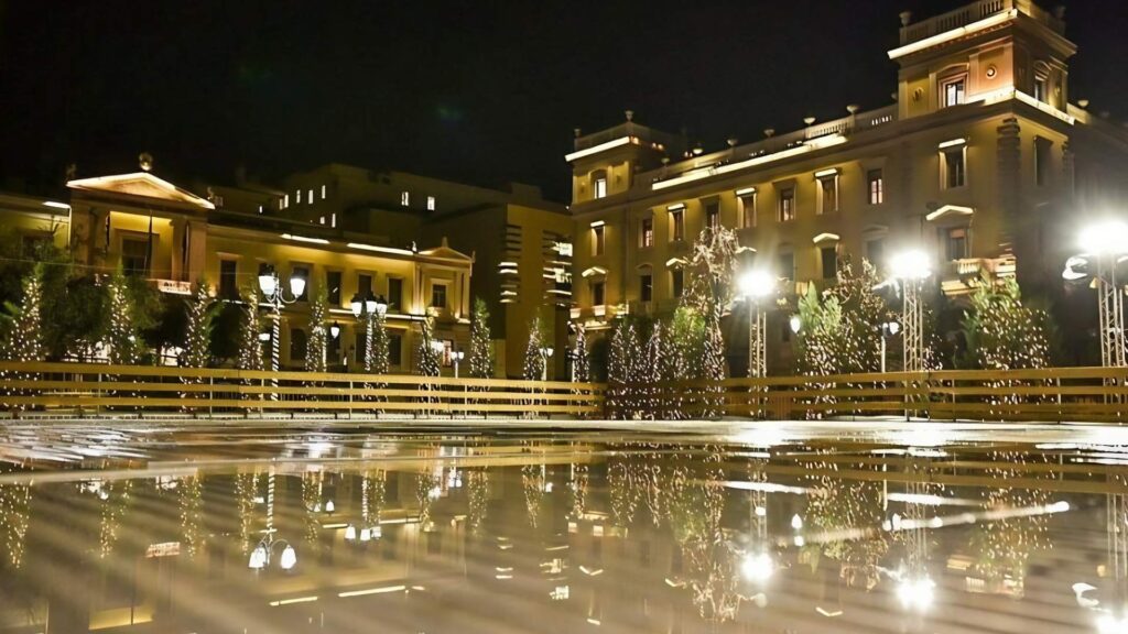 The illuminated buildings and trees in Athens create a stunning reflection on the wet surface at night, offering a magical glimpse into the enchanting things to do during an Athens Christmas.