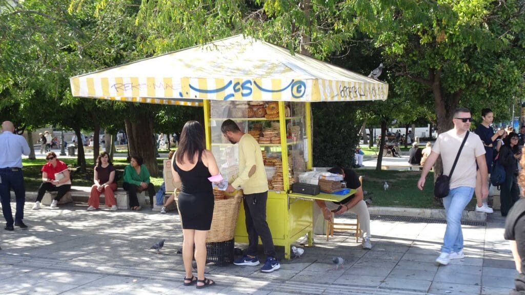 People are buying Greek street food from a small yellow kiosk brimming with snacks. Trees and pedestrians create a lively backdrop, enhancing the scene's charm.