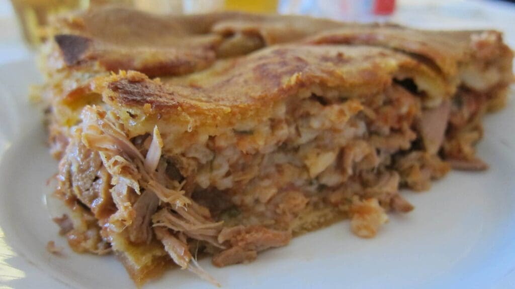 A slice of pie with a filling composed of shredded meat and rice offers a delightful nod to Greek street food, served elegantly on a white plate.