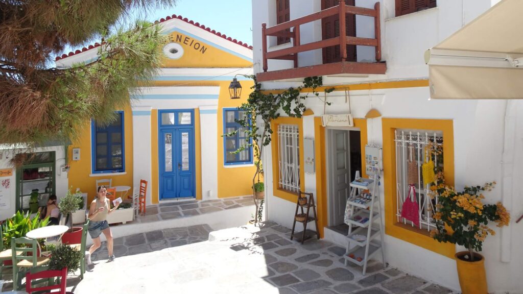 A charming courtyard in Naxos features a white and yellow building, bright blue doors, and a small café. Potted plants and a few tables with chairs decorate the area.