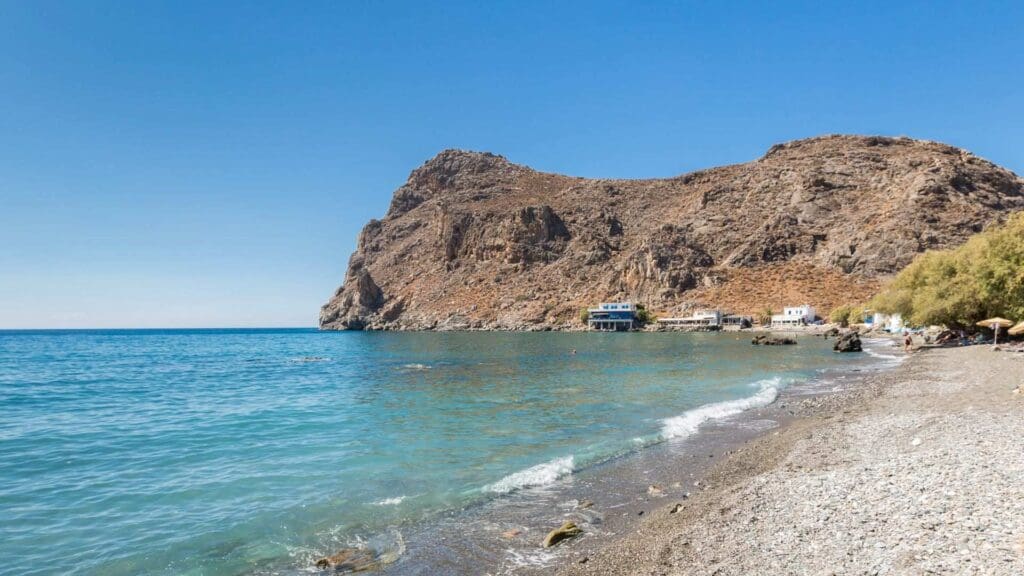 Rocky beach with clear turquoise water, small waves, and arid hillside in the background. A few buildings near Heraklion beaches line the shoreline under a clear blue sky.