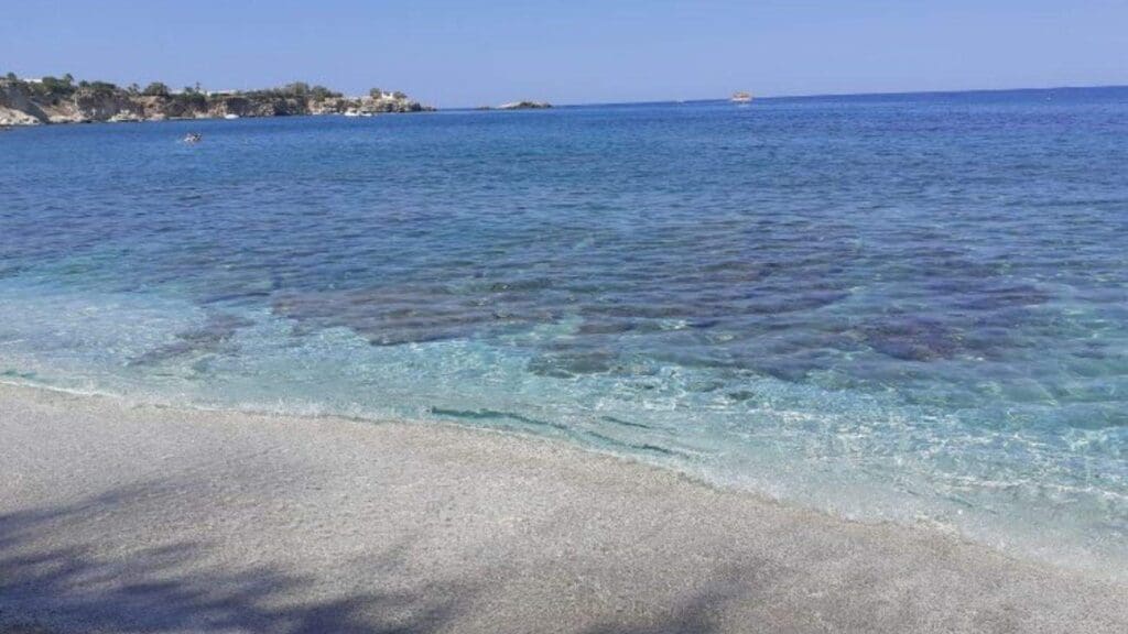 A serene Heraklion beach with clear blue water and gentle waves lapping against the sandy shore under a clear sky, where distant land is visible on the horizon.