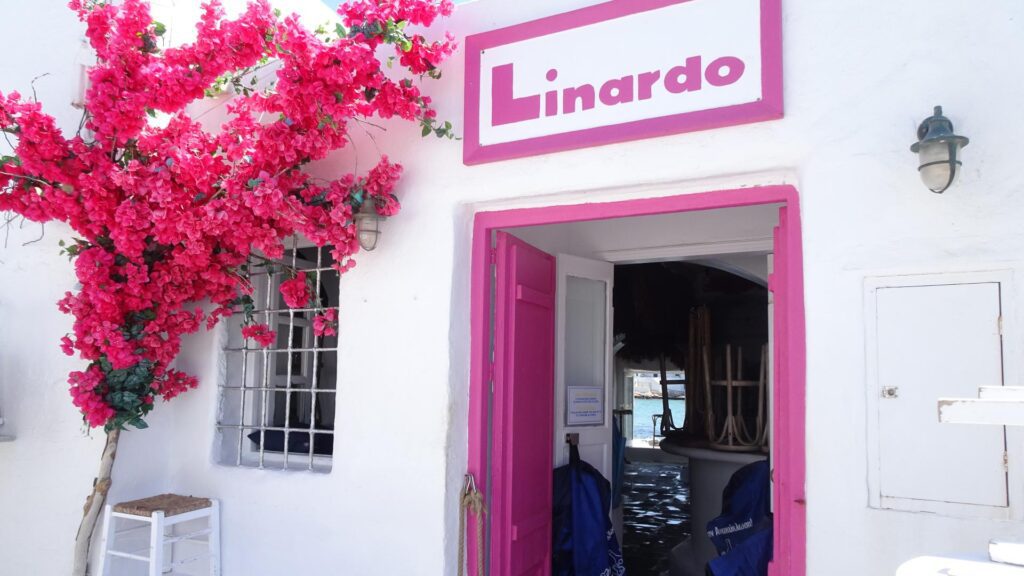 A white building on Milos boasts a pink sign reading "Linardo." Pink flowers decorate the entrance, and a single door is open, offering a glimpse inside this charming spot.
