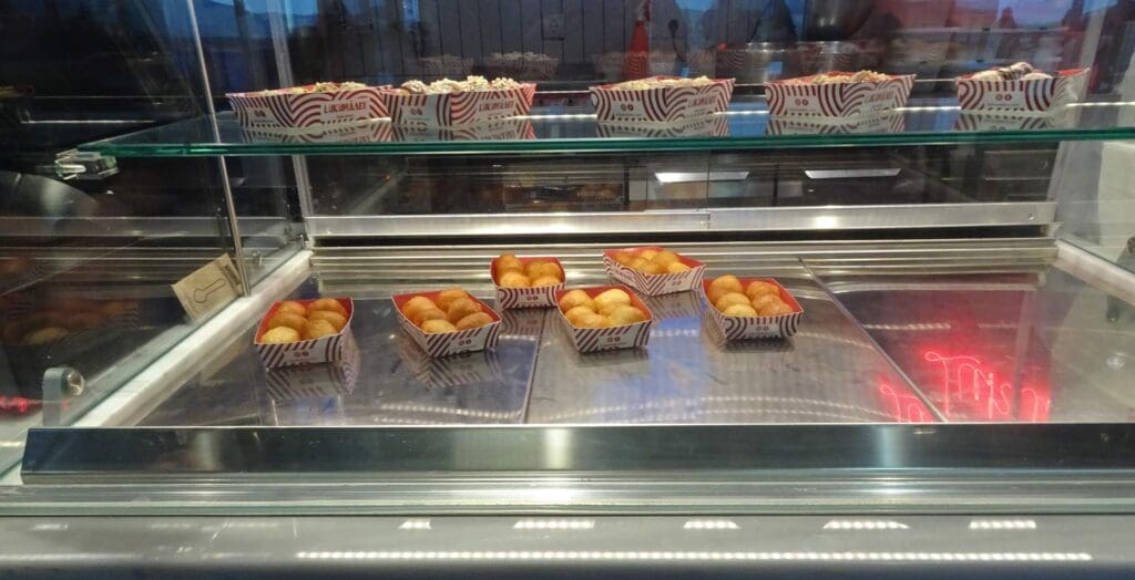 Trays of round fried snacks, reminiscent of Greek street food, and popcorn in striped boxes rest on a stainless steel counter under a glass display.