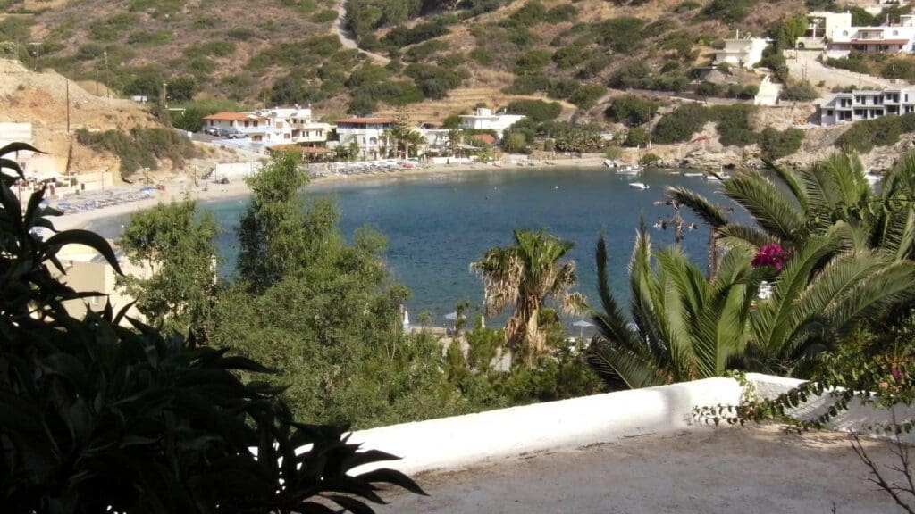 Bay view with clear water, surrounded by lush greenery and hillside houses reminiscent of Heraklion beaches. A few small boats gently glide across the water.