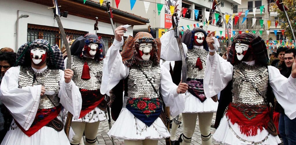 Five people in traditional Greek costumes with white masks and metallic chest armor stand in a festive street setting adorned with colorful flags, embodying the vibrant spirit of Apokries.