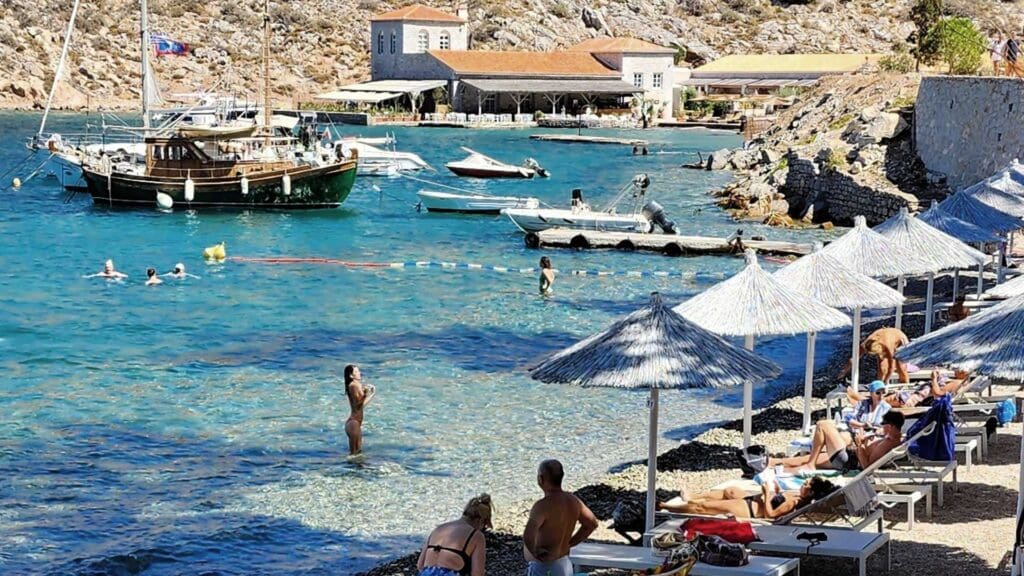 On the enchanting Hydra beaches, people sunbathe on loungers under colorful umbrellas by a rocky shore. Several boats gracefully float in the turquoise water nearby, with charming buildings serving as a picturesque backdrop.