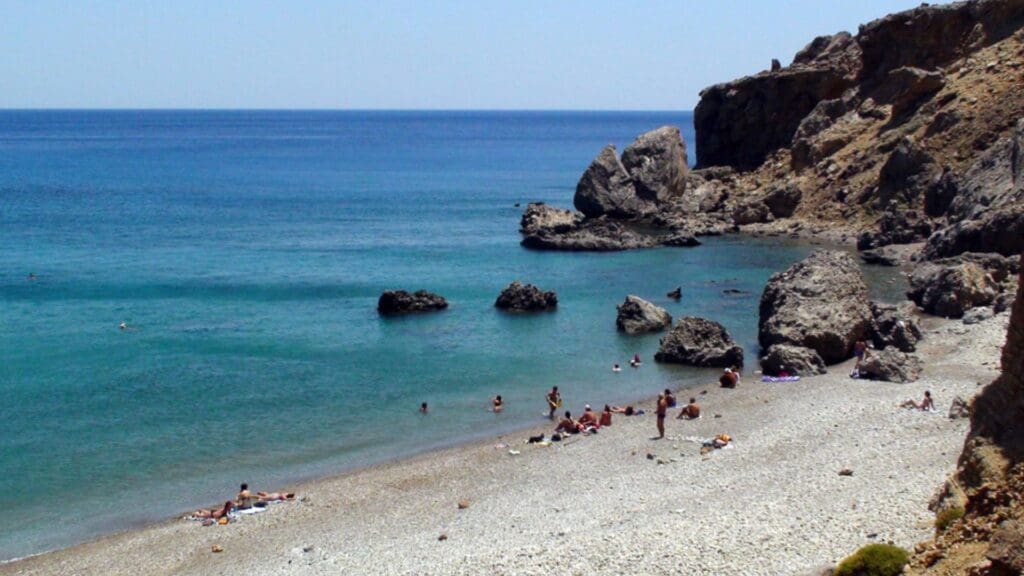 A rocky beach with crystal-clear water, people sunbathing and swimming, bordered by steep cliffs under a clear blue sky reminiscent of Heraklion beaches.