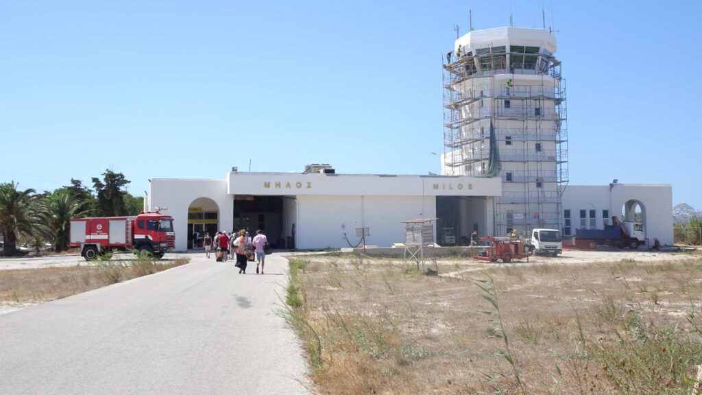 People walk toward a small, under-renovation airport terminal with a scaffolding-covered control tower, reminiscent of the charming simplicity found in island guides for places like Paros.