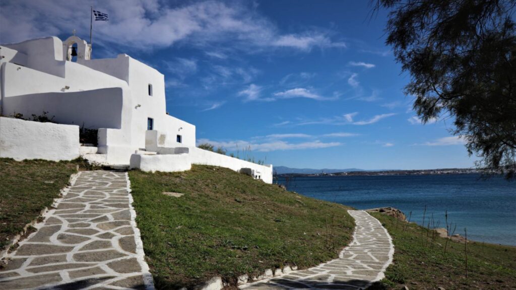 White building in Naoussa, Paros, with a Greek flag fluttering near the sea, a stone pathway inviting you to explore. Tree branches frame the scene against a backdrop of endless blue sky.